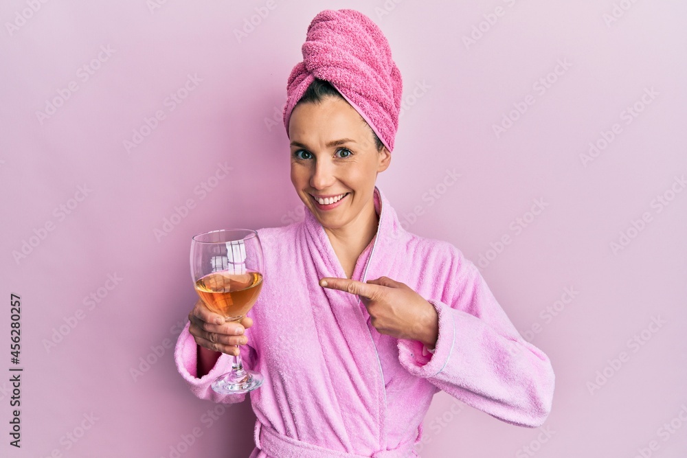 Young blonde woman wearing bathrobe drinking a glass of white wine smiling happy pointing with hand and finger Young blonde woman wearing bathrobe drinking a glass of white wine smiling happy pointing with hand and finger