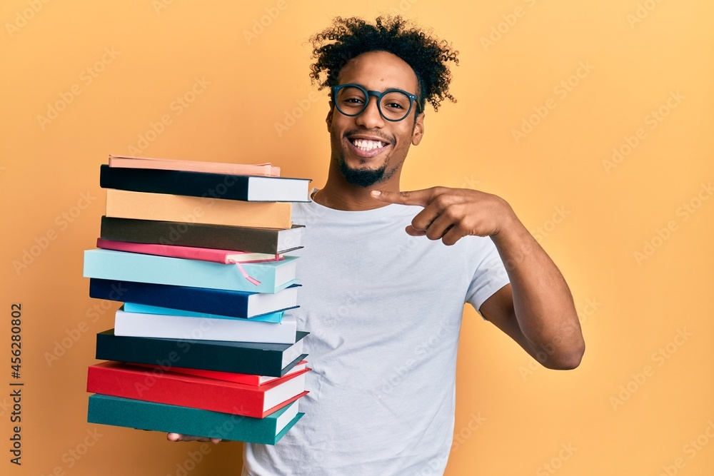 Young african american man with beard holding a pile of books smiling happy pointing with hand and finger Young african american man with beard holding a pile of books smiling happy pointing with hand and finger
