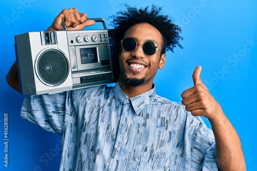 Young african american man with beard holding boombox, listening to music smiling happy and positive, thumb up doing excellent and approval sign
