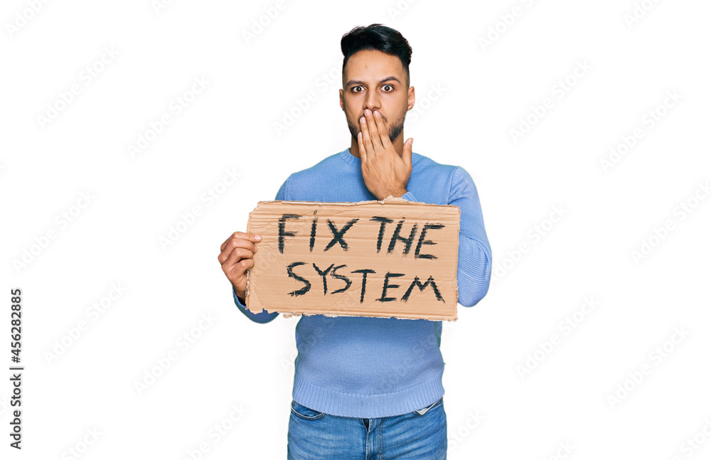 Young arab man holding fix the system banner cardboard covering mouth with hand, shocked and afraid for mistake. surprised expression Young arab man holding fix the system banner cardboard covering mouth with hand, shocked and afraid for mistake. surprised expression