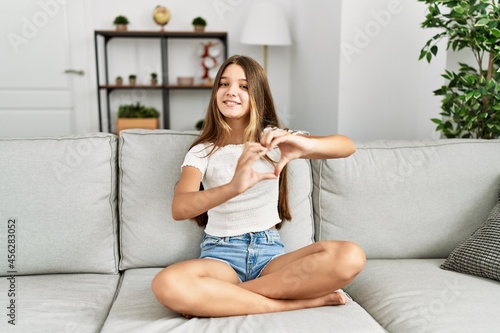 Young brunette teenager sitting on the sofa at home smiling in love doing heart symbol shape with hands. romantic concept.