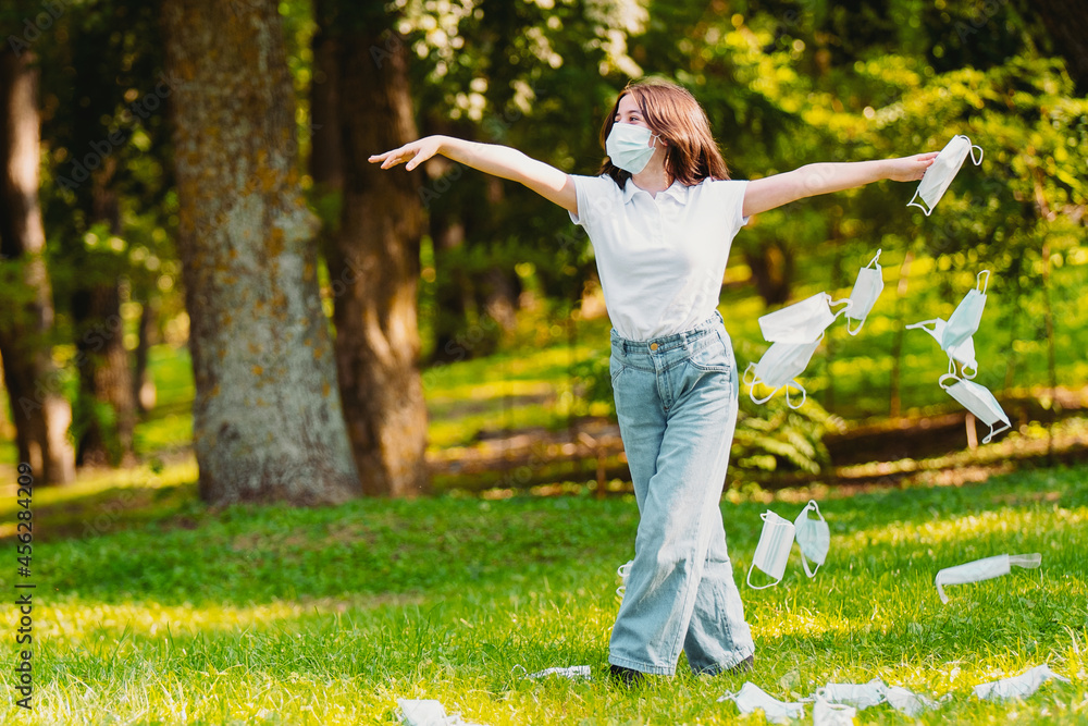 Full-length photo of a young woman wearing a facial medical mask holding hands by the sides and throwing medical mask around her outdoors. Full-length photo of a young woman wearing a facial medical mask holding hands by the sides and throwing medical mask around her outdoors.