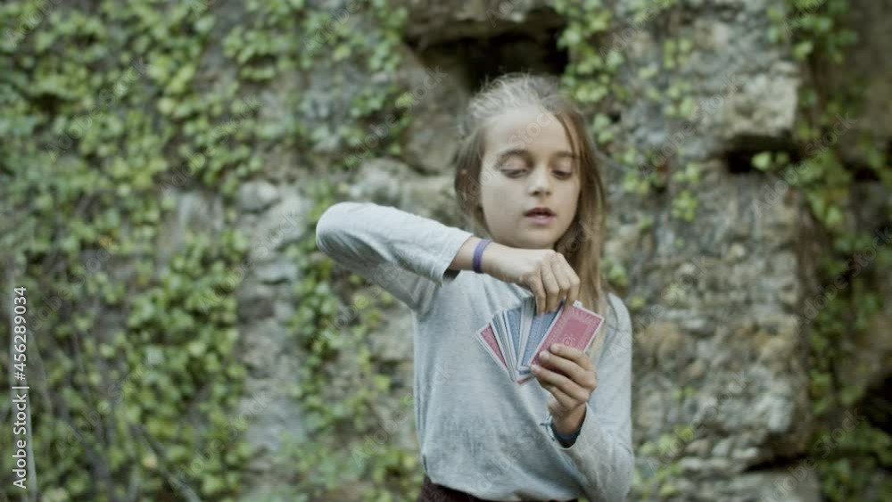 Funny girl standing among walls of old castle and reading cards. Beautiful Caucasian teenager pretending to be witch, gesticulating, drawing card from pack and smiling. Magic, witchcraft concept Funny girl standing among walls of old castle and reading cards. Beautiful Caucasian teenager pretending to be witch, gesticulating, drawing card from pack and smiling. Magic, witchcraft concept