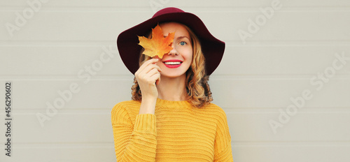 Autumn portrait of beautiful happy smiling young woman covering her eyes with yellow maple leaves wearing a knitted sweater, hat on gray background