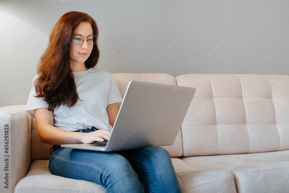 A young woman works at home behind a laptop sitting on a beige couch. The concept of blogging and home learning. A young woman works at home behind a laptop sitting on a beige couch. The concept of blogging and home learning.