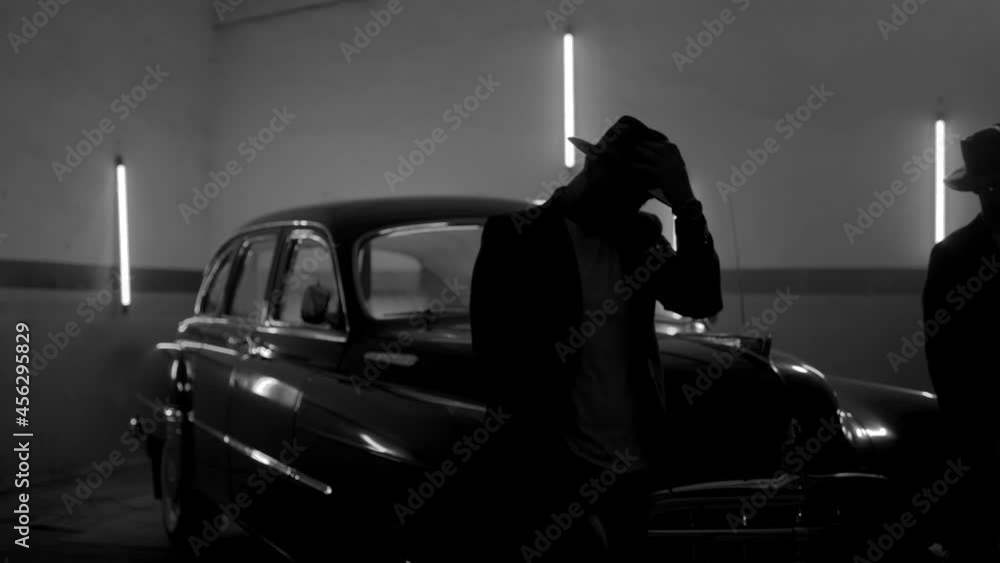 Two silhouettes of men in black suits with classic hats standing by vintage old car. Strangers look like italian mafioso from movie . Footage have permissible count of normally film effect style GRAIN