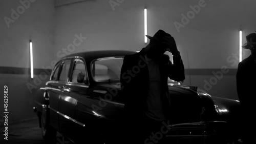 Two silhouettes of men in black suits with classic hats standing by vintage old car. Strangers look like italian mafioso from movie . Footage have permissible count of normally film effect style GRAIN photo