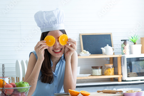 Asian woman wears chef hat .cover her eyes with slice of orange fruit, playing with someone, in the kitchen at home
