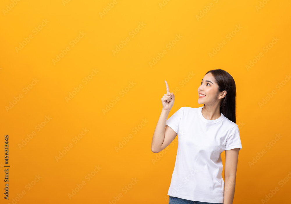 Beautiful asian woman pointing finger into blank space. Smiling female wearing white shirt on isolated yellow background. Attractive Teenager girl looking at empty space for text. Beautiful asian woman pointing finger into blank space. Smiling female wearing white shirt on isolated yellow background. Attractive Teenager girl looking at empty space for text.