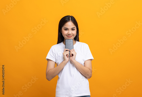 Pretty young asian woman using smartphone standing on isolated yellow background feeling happy. Shopping online payment with mobile phone. Excited and surprised female holding cellphone.