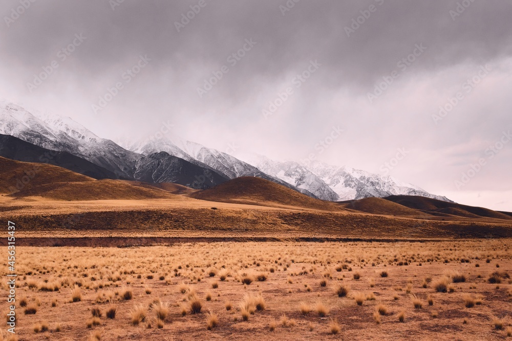 Dry grassland with rolling hills by the snowy Andes mountains in Valle de Uco, Mendoza, Argentina, in a dark cloudy day.