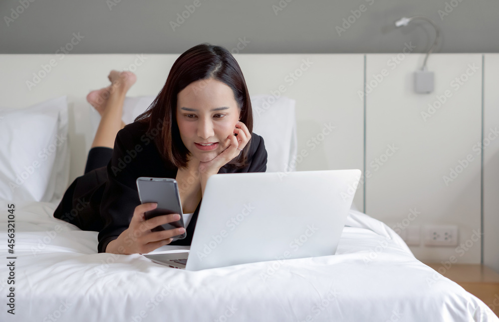 Young asian woman in black suit reading text message on mobile phone while work in the morning in bed with laptop computer. Young asian woman in black suit reading text message on mobile phone while work in the morning in bed with laptop computer.