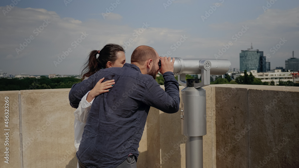 Caucasian tourist looking at panoramic view of metropolis using telescope on sky tower top. Young woman at observation point watching city and nature in metropolitan landscape
