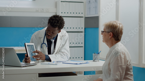 Medic of african american ethnicity holding tablet with chiropractic injury explaining disease to elderly woman with osteoporosis. Black doctor and aged patient looking at screen
