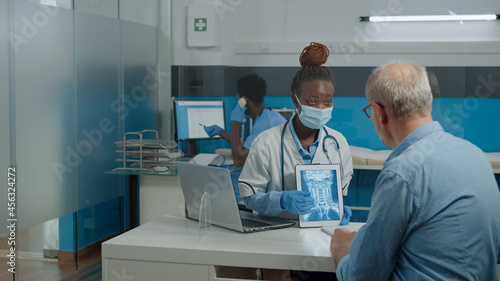 Young doctor analyzing x ray on digital tablet for old patient with disease sitting at desk with plexiglass wall. Medic wearing face mask, explaining radiography to senior man