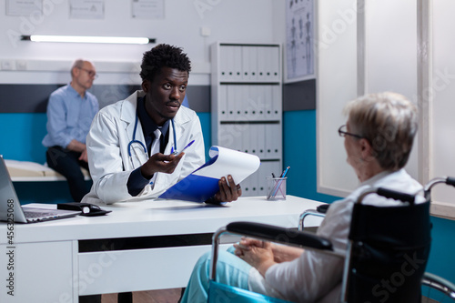 African american medic explaining diagnosis to old disabled woman at medical cabinet. Black man with doctor profession consulting senior patient with handicap sitting in wheelchair