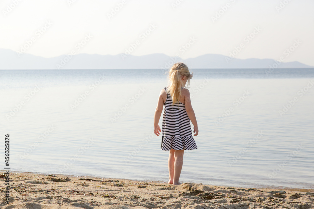 Adorable toddler girl in striped dress enjoying a day at the beach. Idyllic summer vacation.