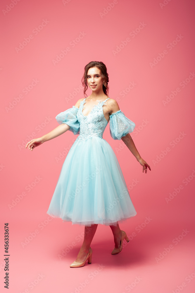 full-length portrait in motion, walking, smiling young woman in blue evening dress with full skirt and lantern sleeves, isolated on pink background. full-length portrait in motion, walking, smiling young woman in blue evening dress with full skirt and lantern sleeves, isolated on pink background.