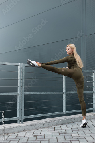 Beautiful young woman athlete with sporty slim body and sexy legs in fashion sportswear with sneakers doing exercises stretching and warm up in the city