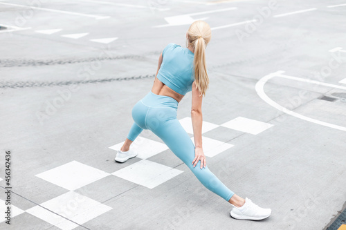 Fitness model woman with blonde hair in blue sporty clothes with white sneakers doing exercise and warm up on asphalt on the street, back view © alones