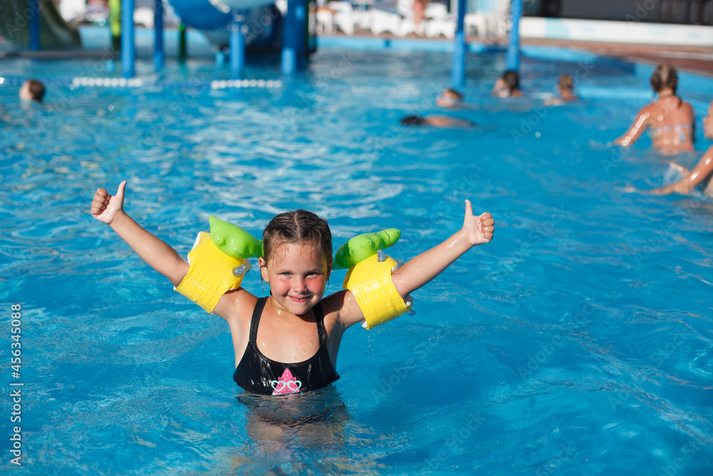 Child poses in water. Happy little girl in floaties raises her hands up and enjoys her vacation at water park. 