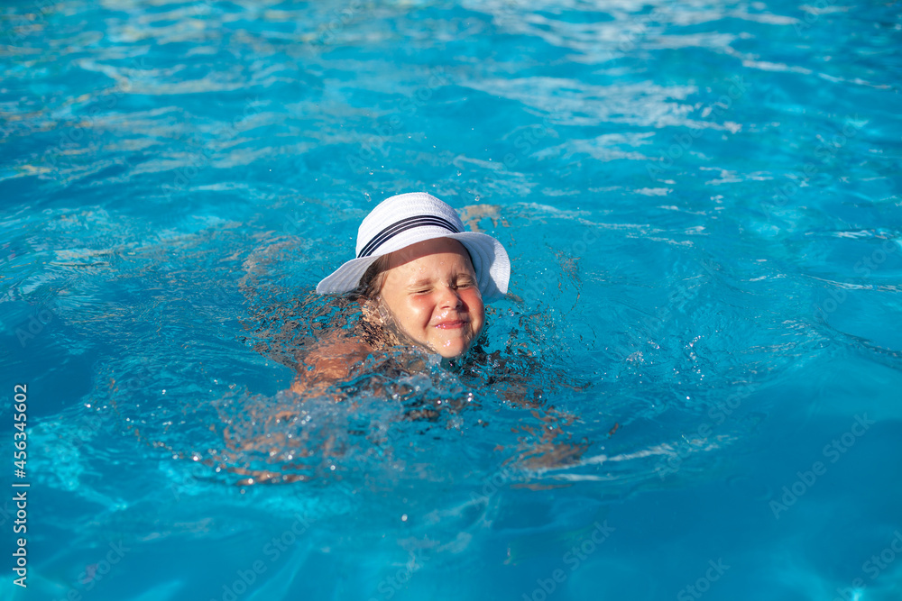 Child in white straw hat is playing in water. Happy charming girl takes swimming lessons and squints from bright summer sun. 