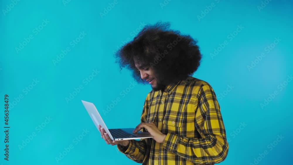 A young man with an African hairstyle on a blue background is typing on a laptop. On a colored background A young man with an African hairstyle on a blue background is typing on a laptop. On a colored background