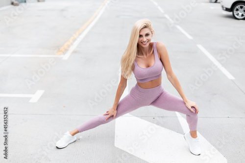 Sporty happy young woman with a smile in stylish sportswear with white sneakers doing stretching exercise and warm up on the asphalt in the street