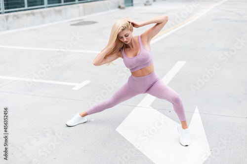 Happy beautiful young woman model in fashion purple sportswear with white sneakers doing stretching exercise in the city on parking lot