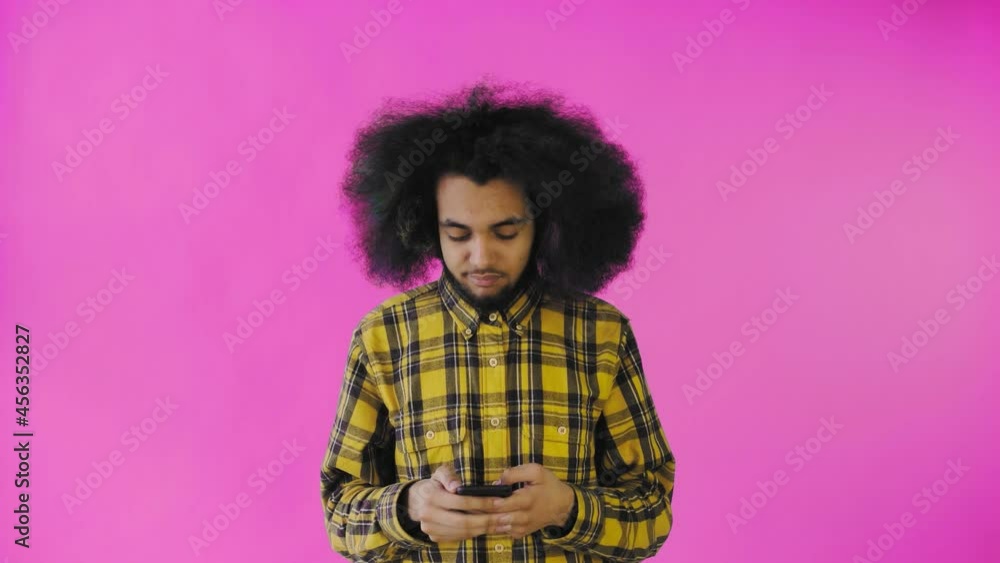A young man with an African hairstyle on a pink background is talking into his phone. A young man with an African hairstyle on a pink background is talking into his phone.