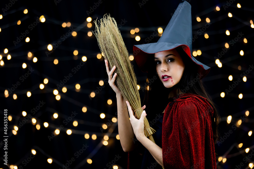 Studio shot of Caucasian female model in black witch costume with tall hat and red shawl holding broomstick play trick or treat in Halloween party traditional event on light bulb bokeh background Studio shot of Caucasian female model in black witch costume with tall hat and red shawl holding broomstick play trick or treat in Halloween party traditional event on light bulb bokeh background