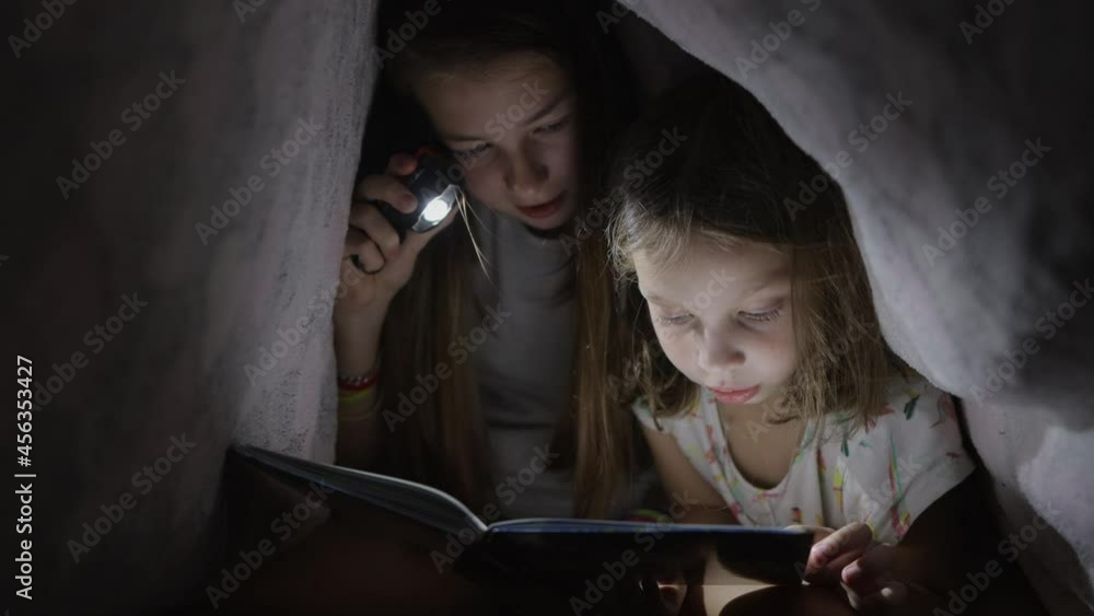two sisters reading a book at night with a flashlight under the covers, vertical video. two sisters reading a book at night with a flashlight under the covers, vertical video.