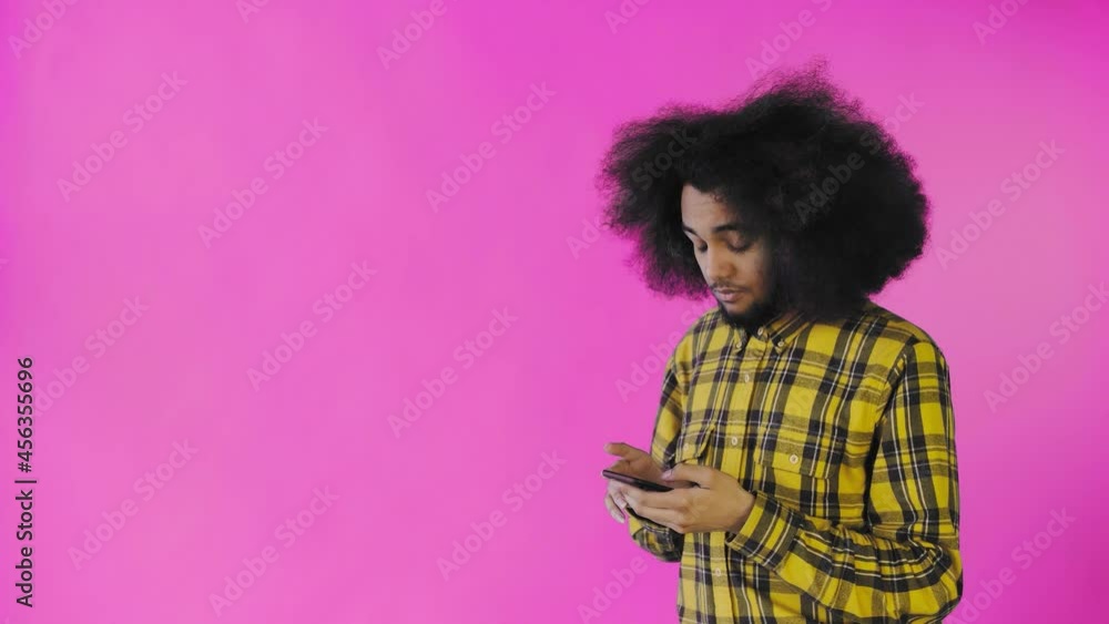 A young man with an African hairstyle on a pink background takes out his phone, looks at the message and puts it back in his pocket. Emotions on a colored background A young man with an African hairstyle on a pink background takes out his phone, looks at the message and puts it back in his pocket. Emotions on a colored background