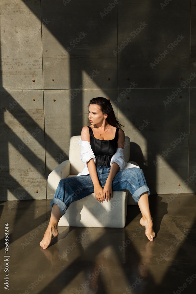 Pretty young woman with big breast posing on white chair in studio, steel wall behind, wearing jeans and white shirt, natural warm sunset light