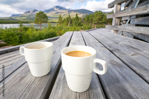 Two white cups filled with coffee with milk on a wooden table with arctic landscape of Swedish Lapland in the background