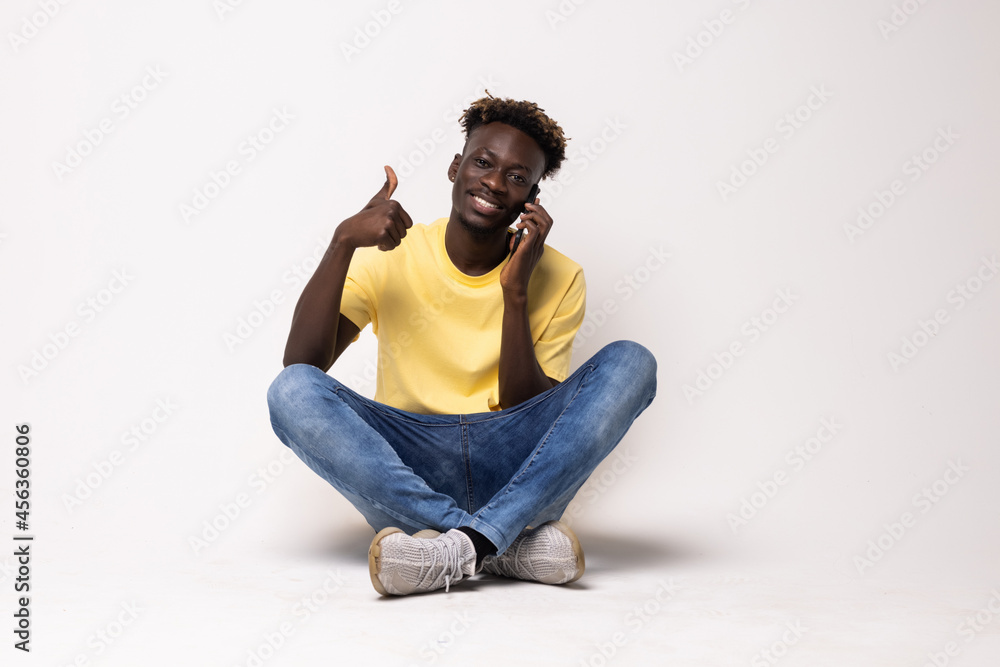 Portrait of a smiling afro american man sitting on the floor with thumbs up and talking on the phone isolated on a white background Portrait of a smiling afro american man sitting on the floor with thumbs up and talking on the phone isolated on a white background
