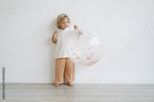 Three years old caucasian blonde boy dressed in white and nude urban clothes holding a big confetti balloon in a white background photo