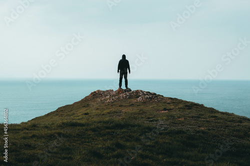 A lone hooded figure standing on top of a hill  looking out across the ocean.