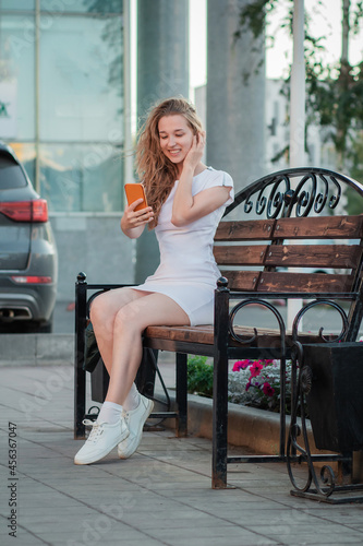 A girl in a simple white dress holds a phone against the backdrop of an office building. Young and stylish woman with a smartphone in the city makes a selfie.
