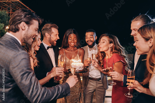 Beautiful young woman in evening gown blowing candles on cake while celebrating birthday with friends