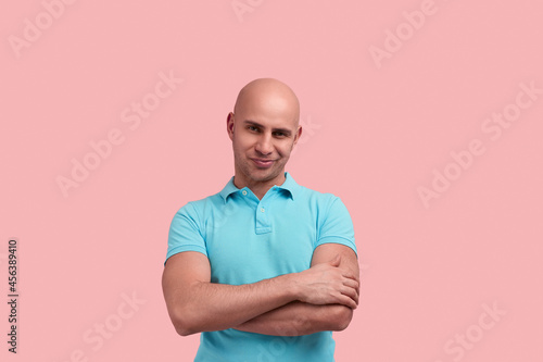 Skeptical bald homosexual man with bristle crosses hands over chest, does not believe information, makes wry mouth, inclines head aside, gay friendly, wears blue polo shirt, poses on pink background.