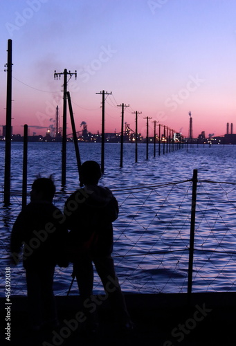 日本の海岸 海の中に在る電柱群と工場群のシルエットを眺める二人の子供のシルエット