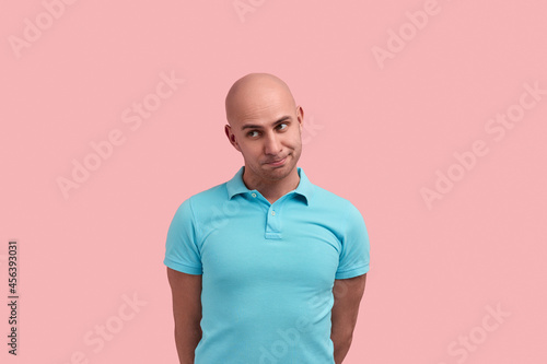 Shy bald homosexual man with bristle is apologising, begs pardon, admits guilt, purses lip and looks sullenly aside , gay friendly, wears blue polo shirt, poses over pink background. photo