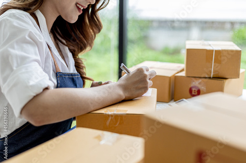Asian woman writing customer's shipping information on parcel box, she owns an online store, she ships products to customers through a private courier company. Online selling concept. © kamiphotos