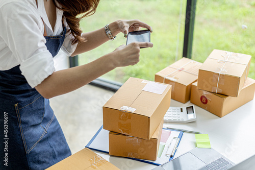 An Asian woman is using a smartphone to take pictures in front of a parcel box for evidence, she owns an online store, she packs and ships through a private shipping company. Online selling concept.