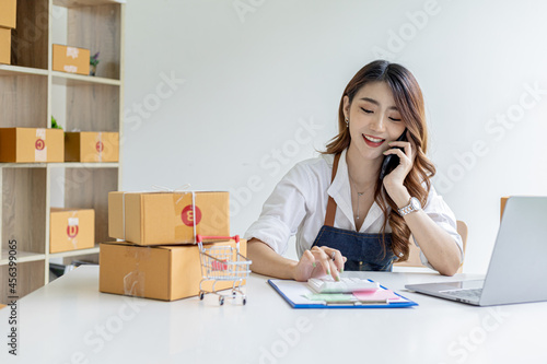 Asian woman is talking on the phone with a customer to confirm an order, she owns an online store, she packs and ships through a private transport company. Online selling and online shopping concepts.
