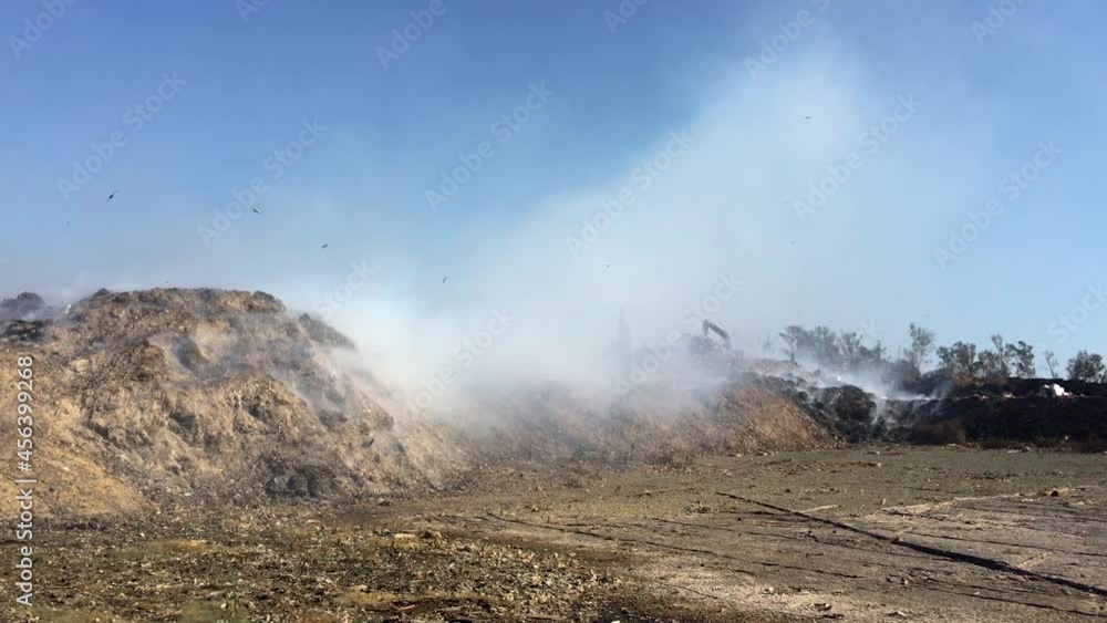 Burning pile of garbage at dump ground or landfill releasing toxic smoke in environment and polluting air. Excavator or earth mover moving or segregating garbage. Burning pile of garbage at dump ground or landfill releasing toxic smoke in environment and polluting air. Excavator or earth mover moving or segregating garbage.