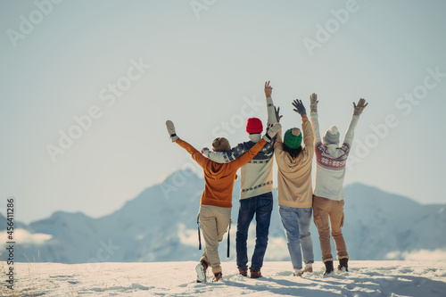 Four happy stylish friends stands, embrace and looks at snow capped mountains. Winter vacations concept photo