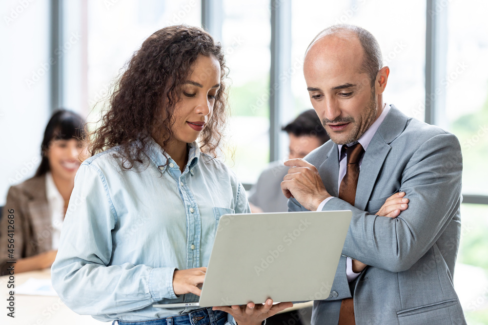 Businessman and businesswoman sharing laptop while standing in office building room. Businessman and businesswoman sharing laptop while standing in office building room.