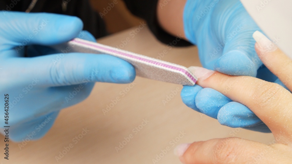 Manicurist files the shape of the nails with a special nail file, close up view. The manicurist shapes the nails with a nail file. The process of nail care in a beauty salon. Nail file processing. Manicurist files the shape of the nails with a special nail file, close up view. The manicurist shapes the nails with a nail file. The process of nail care in a beauty salon. Nail file processing.
